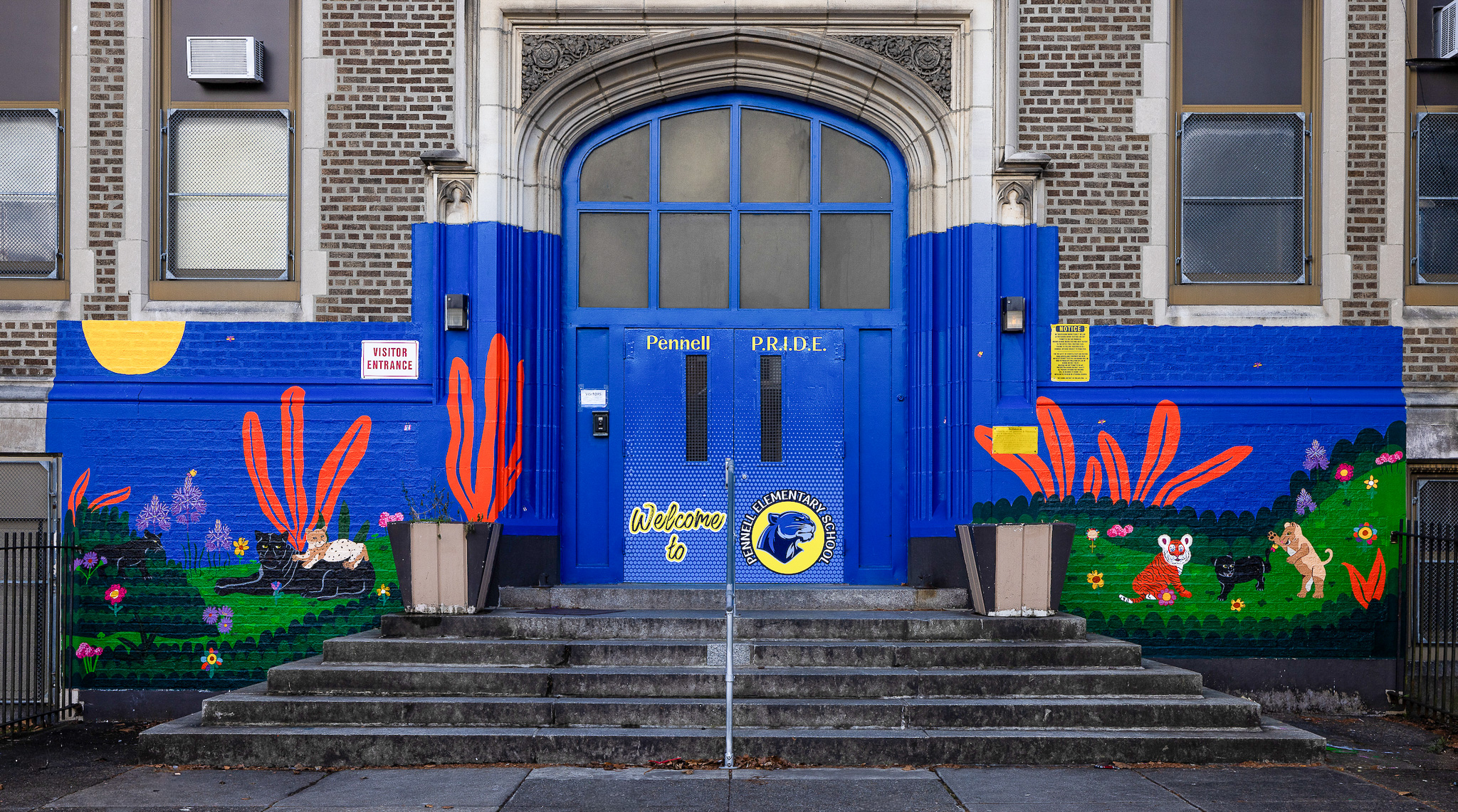 Front entrance of Joseph Pennell Elementary School featuring bright blue doors with the school logo, colorful panther-themed mural artwork, landscaped planters, and a welcome sign at the main entrance.